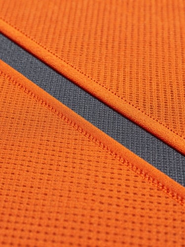 Macro shot of high-contrast orange and charcoal sublimated athletic fabric under stadium floodlights, showing precise stitch lines and vibrant mesh texture.