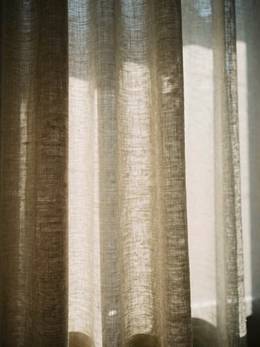 Warm editorial photograph, dramatic window light casting sharp diagonal shadows across a textured linen blackout curtain, shallow depth of field, 35mm