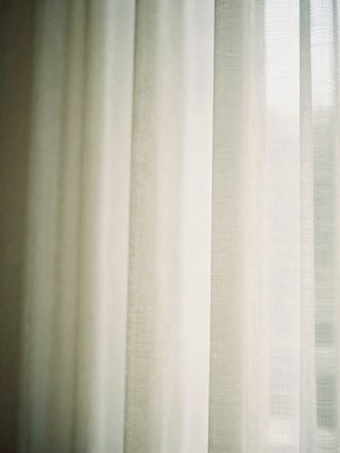 Soft architectural detail showing gentle morning light entering a quiet room through a sheer curtain, warm cream and sage green tones, shallow depth of field, 35mm