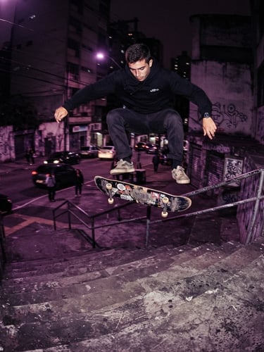Dynamic low-angle action shot of a skateboarder performing a trick over concrete stairs in São Paulo at night, dramatic purple and magenta streetlights, gritty urban texture, 35mm film