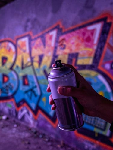 Extreme close-up of spray paint cans held by youth in front of a massive fresh colorful graffiti mural, deep purple overlay, dramatic high-contrast street lighting.