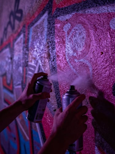 Extreme close-up of hands holding spray paint cans, spraying a fresh graffiti mural on a raw concrete wall, high-contrast night street lighting, deep purple and magenta color grading
