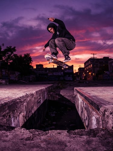 Dynamic low-angle wide shot of a skateboarder performing a trick over a concrete gap at sunset, deep purple and magenta color grading, high contrast street lighting.