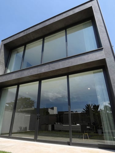 Low-angle architectural photograph of a modern luxury residence facade in Goiânia, expansive floor-to-ceiling glass sliding doors with slim dark aluminum frames reflecting a clear blue sky, bright daylight.