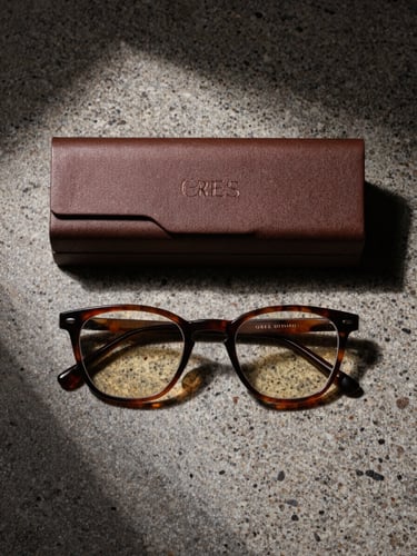 Overhead flat-lay of a complete luxury eyewear set—frame and closed archival box side by side on raw travertine, sharp diagonal shadow cutting across both objects, deep umber tones, controlled studio light from a single high source, no text visible, packaging lid embossed with subtle texture, total product isolation on stone