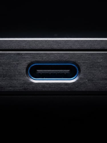 Macro close-up shot of a USB-C port on a dark brushed-metal laptop chassis, sharp studio strobe lighting with crisp electric blue rim highlights, deep obsidian background