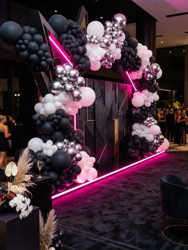 Wide-angle portrait of a luxury event setup, sharp architectural balloon installation in deep charcoal and white with neon pink smart LED lighting glowing from beneath, a modern standing backdrop on a dark custom carpet, luxury editorial photography, 35mm lens