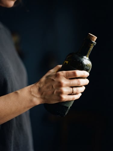 Moody editorial food photography, close-up of a hand inspecting a dark glass bottle of olive oil under soft natural side-light, gold highlights, deep navy background.
