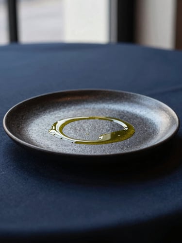 Elegant minimalist restaurant in Copenhagen, soft natural light, a single dark ceramic plate with a delicate drizzle of golden olive oil, deep navy tablecloth, high-end editorial style.