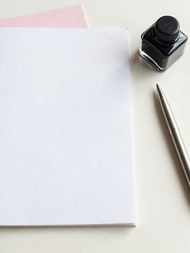 Overhead flat-lay of heavy-weight cotton paper stock, a modern glass ink well, and a single metal pen resting on a clean cream plaster surface under soft morning light.