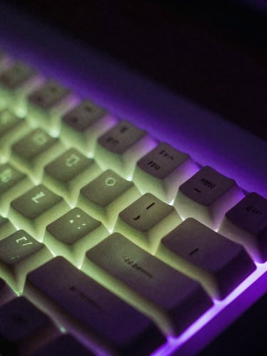 Macro shot of a glowing mechanical keyboard in a pitch-black room, illuminated by a harsh violet screen glow, cinematic film grain