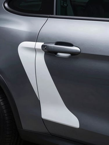 Tall portrait — close study of a vehicle's rear quarter panel where a custom graphic wraps precisely around a compound curve and door handle recess. The vinyl edge is immaculate, following the body crease with surgical accuracy. Cold studio strobe lighting from the upper left sculpts the surface, revealing the graphic as a three-dimensional architectural decision rather than a flat decal. Dark charcoal vehicle surface, white and silver graphic elements. No people, no distracting backgrounds.
