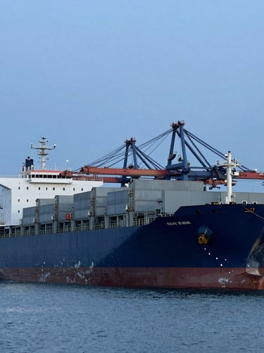 Wide-angle industrial photography of a massive cargo ship docked at a modern port terminal, cool blue-hour daylight, deep navy and steel gray tones, 35mm lens.
