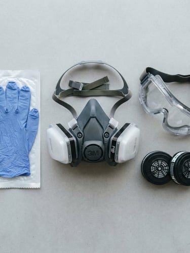 Flat-lay of professional safety gear, including a modern half-mask respirator with dual filters, nitrile gloves, and clear safety goggles, laid out on a clean light gray surface, bright overcast natural light.