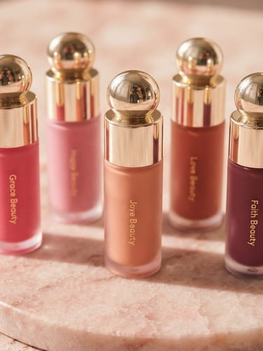 Elegant close-up of Rare Beauty liquid blush bottles on a soft pink stone surface, warm diffused daylight, gold reflections, shallow depth of field, 35mm
