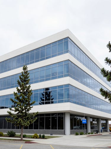 Wide-angle architectural shot of a modern office building exterior in Coeur d'Alene, surrounded by pine trees under bright overcast daylight, clean glass reflections