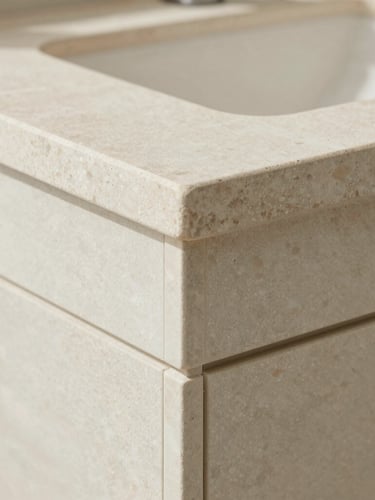 Extreme close-up architectural photograph of a perfectly executed mitered edge joint on a premium stone vanity, bright natural coastal daylight, pale sage tones, crisp detail
