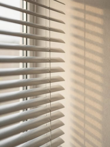 Angled close-up of aluminium venetian blind slats, fine horizontal lines casting parallel shadow bands on a warm cream wall, soft diffused daylight, no people