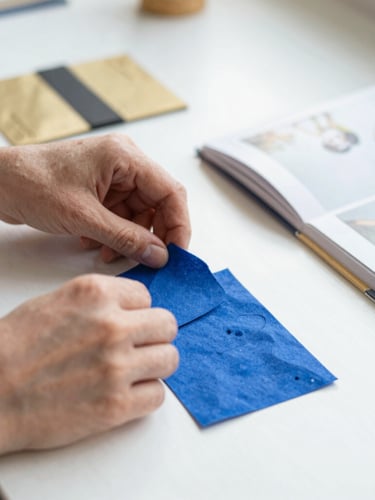 Close-up of hands arranging tactile sewing materials and high-quality printed books on a clean white workspace, soft morning light, shallow depth of field, 35mm