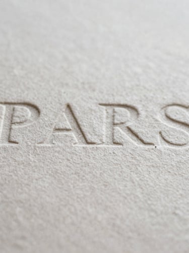 Macro photograph of heavy textured cotton paper with debossed typography, soft shadows, natural overcast Parisian light, editorial film style
