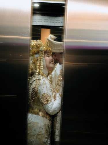 A smiling couple in traditional white and gold wedding attire glimpsed through closing elevator doors.
