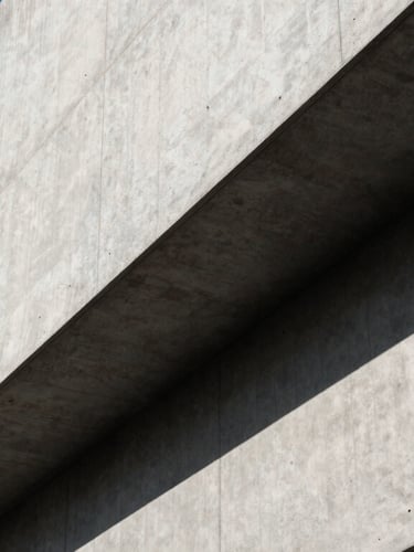 A high-contrast architectural close-up of a modern concrete building facade with sharp diagonal shadows, clean lines, and a sliver of bright blue sky.