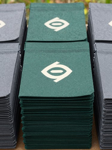 Straight-on product layout of neatly stacked nonwoven bags in deep forest green and slate gray, showing crisp printed brand graphics, bright natural daylight
