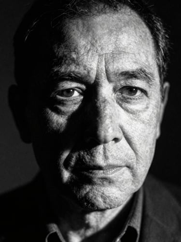 High-contrast black and white portrait of a thoughtful, middle-aged writer with deep-set eyes, looking slightly off-camera. Harsh chiaroscuro lighting, long shadows, grainy film texture, 35mm lens.