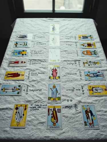Overhead study of a tarot spread laid methodically on a linen cloth, cards face-up with handwritten annotation notes beside them, cool diffused daylight from a studio window, no hands in frame