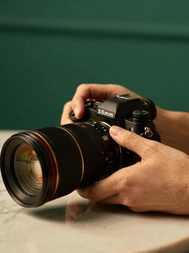 Warm editorial photography of hands adjusting a professional camera lens on a marble table, soft studio daylight, rich emerald and gold tones, 35mm