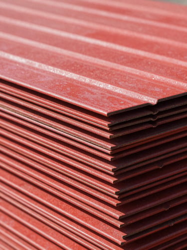 Close-up of corrugated red metal sheets stacked in bright direct daylight, high-contrast metallic textures, sharp focus on the real thickness profile