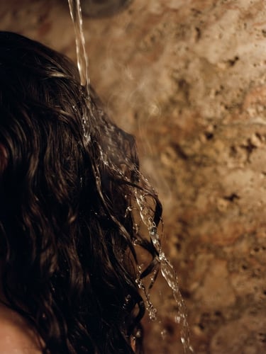 Extreme close-up of warm water slowly flowing over dark hair, steam rising, soft low-key candlelight, travertine stone texture in background, tactile, 35mm