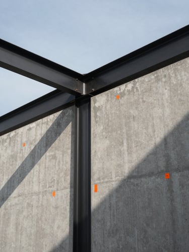 Wide panoramic 21:9 view of a blackened steel beam intersecting a raw concrete wall, sharp safety-orange construction markings visible, heavy geometric shadows cast by low-angle morning sun, generous negative space.