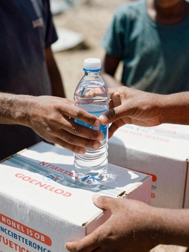 Candid close-up photo of hands distributing water bottles and emergency food boxes to families in a temporary shelter, natural harsh daylight, high contrast