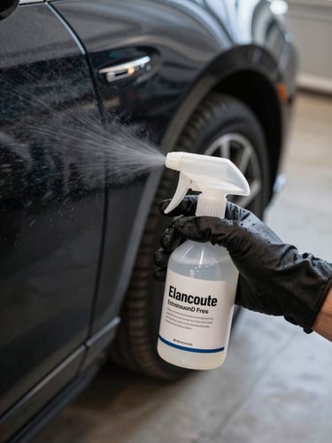 Close-up overhead shot of a gloved hand holding a spray bottle of encapsulant product aimed at a dark car bonnet surface, residential garage floor visible below, natural daylight, fine mist visible in the air