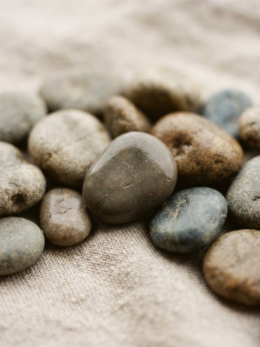 A close-up of smooth, warm river stones resting on textured linen under soft, diffused coastal daylight, shallow depth of field, 35mm.