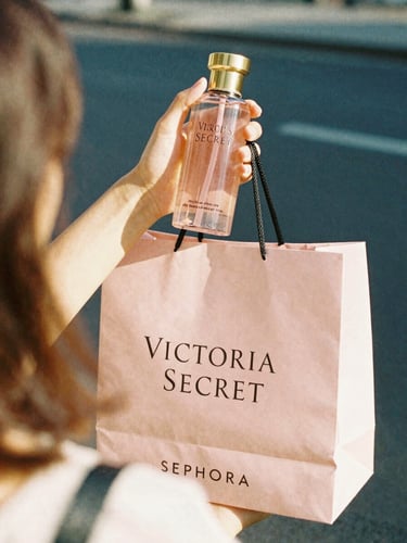 Candid over-the-shoulder shot of a hand holding a Victoria's Secret body mist and a Sephora shopping bag, bright natural daylight, warm golden hour, 35mm film style.