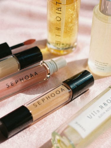 Close-up of viral Sephora lip oils and Bath and Body Works mists arranged on a soft pink satin surface, warm golden hour sunlight, shallow depth of field, 35mm film style