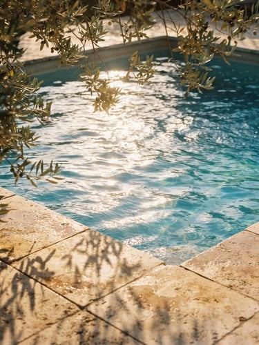 Close-up of warm golden-hour light rippling across a heated private pool, olive leaves casting soft shadows on sun-bleached stone tiles, 35mm film aesthetic.