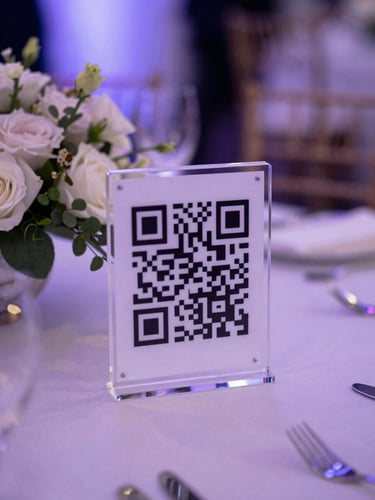 A sleek acrylic QR code stand sitting on a beautifully decorated wedding table next to a floral centerpiece, soft violet lighting, shallow depth of field.