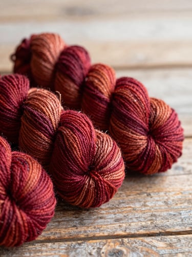 Macro photograph of rich crimson and ochre hand-dyed yarn skeins resting on a rustic wooden table, soft side daylight, shallow depth of field