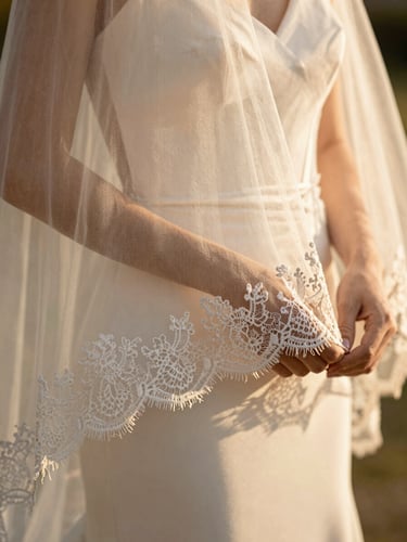 Close-up of a bride's hands holding a delicate lace veil in warm golden-hour sunlight, soft champagne tones, shallow depth of field