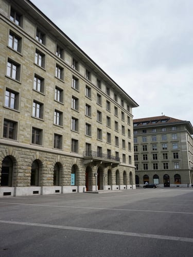 Wide architectural exterior of a Geneva financial quarter building at midday, pale overcast Swiss sky, stone facade detail in the left third, open plaza receding to the right, no figures