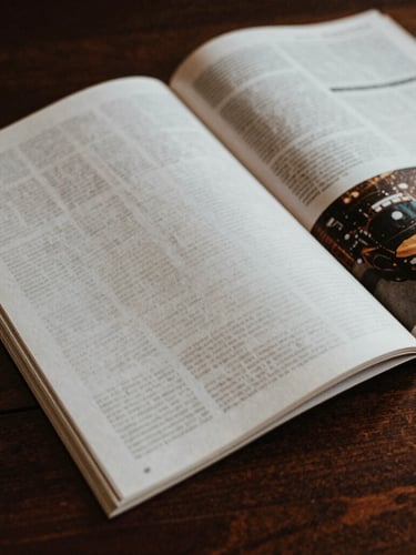 Close-up of tactile printed magazine pages on a dark wooden desk, natural high-contrast side lighting, deep rich tones, 35mm film style.