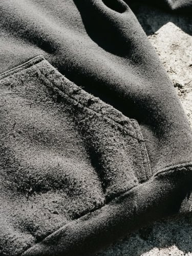 Close-up high-contrast streetwear photography of a heavyweight charcoal hoodie texture, harsh direct sunlight casting sharp shadows on concrete, desaturated tones, 35mm lens