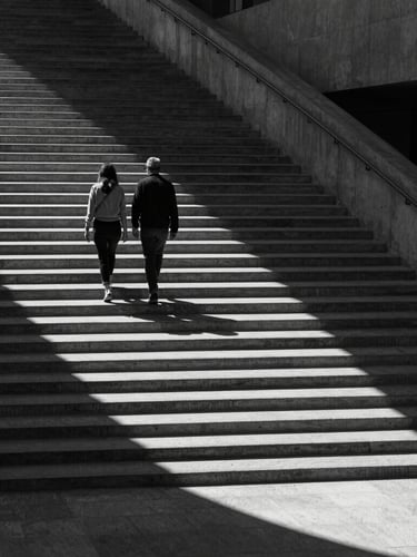 Wide cinematic environmental shot of a couple walking down a stark concrete staircase, dramatic high-contrast natural light casting long shadows, sharp monochrome, architectural framing.