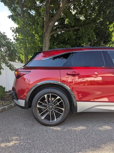 Side profile of a red Acura MDX luxury SUV parked on a paved driveway under a leafy green tree.