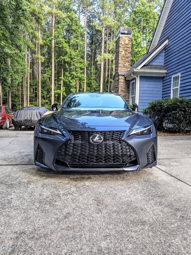 Front view of a charcoal gray Lexus luxury sedan parked on a concrete driveway in front of a blue house.