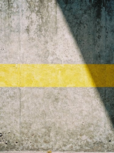 Gritty editorial photography, high-contrast natural light, desaturated urban tones, sharp 35mm film look. Close-up of a weathered concrete wall in San Luis Potosí, marked with a bright industrial yellow stripe, sharp diagonal midday shadows.