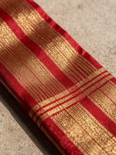 Extreme close-up macro-photography of a Patan Patola saree selvage, showing the mathematical precision of the double-ikat weave and the organic sheen of pure silk. Soft, directional daylight catches the crimson and gold threads against a background of raw plaster.